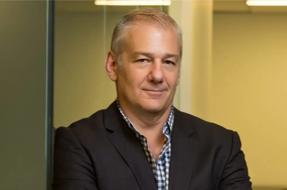 Jeffrey Finkle — silver-haired, dark blazer over a checked shirt, warm half-smile, in a glass-walled New York office